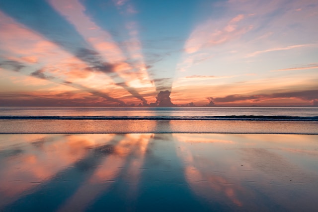 An image of a beach at sunset, the rays of the sun are split by couds in the distance into beams of orange and yellow light. The water is dead calm, reflecting the sky.